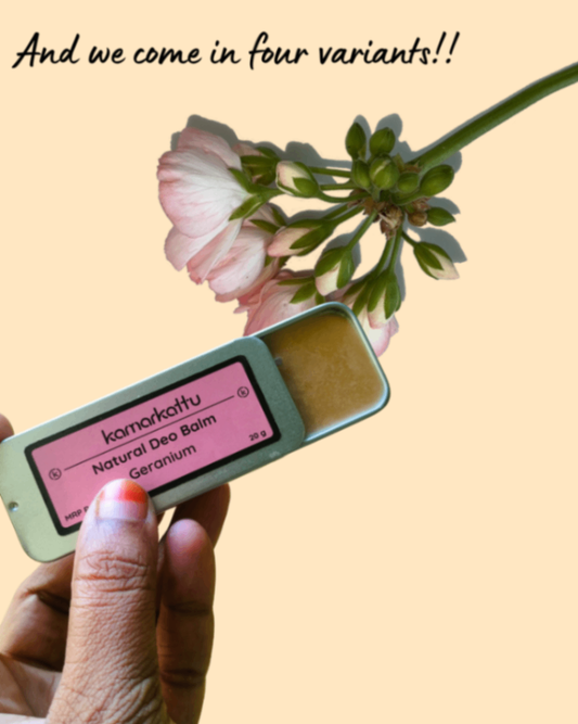 Hand holding an open Kamarkattu deodorant balm tin with a pink Geranium label beside pink flowers.